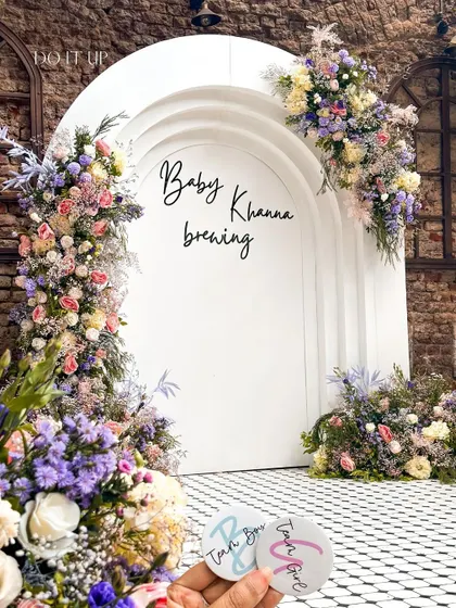 A stunning floral archway for a baby shower. The white arched backdrop, which reads 'Baby Khanna brewing', is adorned with an abundance of pastel flowers, creating a perfect photo opportunity and entrance piece.