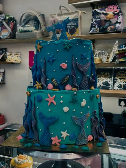A two-tiered mermaid cake displayed in my shop. The deep teal and blue colors create a rich underwater scene, decorated with various sea life.