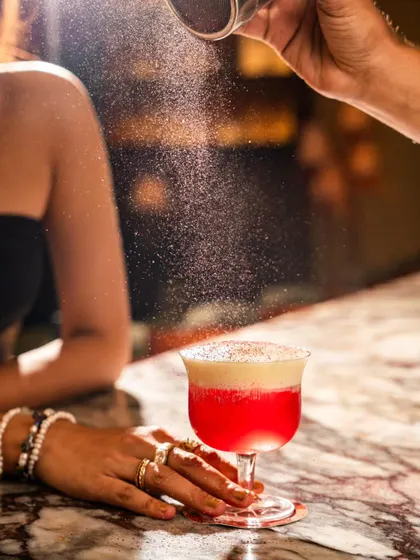 An action shot of a bartender dusting a pink cocktail with a powder garnish at Lyla Bombay. The captured motion of the dust adds a magical quality to the image.