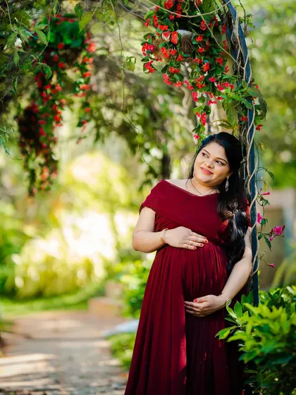 A quiet, contemplative portrait of an expectant mother in a deep red off-the-shoulder gown. The natural frame of the flowering vine adds a touch of romance.
