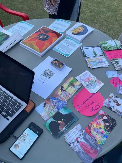 A flat lay of the items on my table at the Youth Alliance event. You can see my laptop for payments, a QR code, and various art prints and cards ready for people to browse. It shows the practical side of being an independent artist.