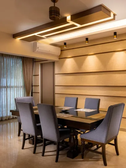 This dining area showcases a custom wooden false ceiling with integrated lighting, creating a warm and focused ambiance. The textured accent wall and upholstered chairs add layers of comfort and style.