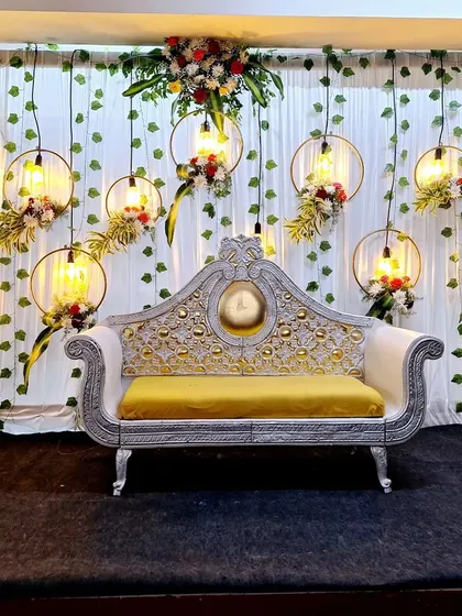 An elegant reception stage design with a classic white and gold loveseat. The backdrop features hanging floral hoops with vintage style bulbs, creating a warm and romantic ambiance.