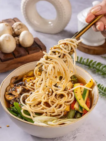 A comforting bowl of noodle soup with fresh vegetables, with a noodle pull shot that makes it look extra appetizing.