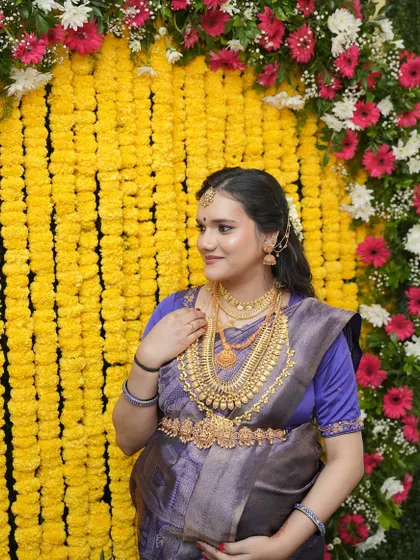 A side profile of the mom-to-be, Varsha, against a vibrant floral backdrop. Her makeup is kept soft to complement her traditional attire and heavy gold jewelry.