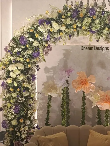 A close-up of a beautiful floral arch designed for a baby shower. The mix of white, yellow, and purple flowers is complemented by whimsical, oversized paper flowers in the background.