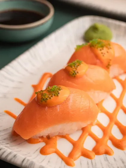 A close-up of salmon nigiri, beautifully plated with a spicy mayo drizzle. The tight crop focuses on the freshness of the fish and the vibrant colors.