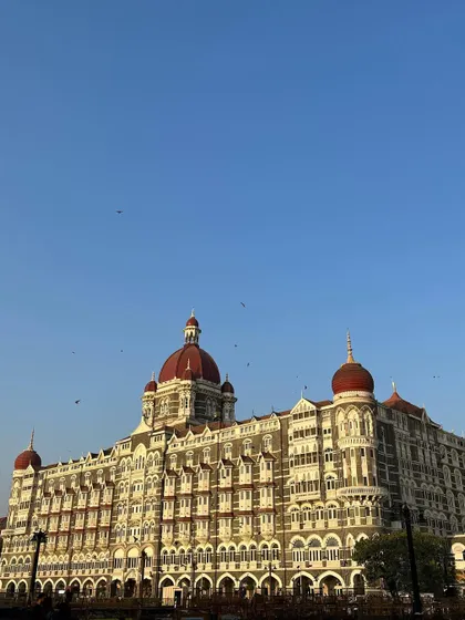 The heritage wing of the Taj Mahal Palace hotel under a clear blue sky, with birds flying around its grand facade.