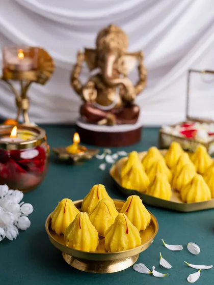Bright yellow kesar modaks offered during Ganesh Chaturthi, with a blurred Ganesha idol in the background creating a sense of depth and focus.