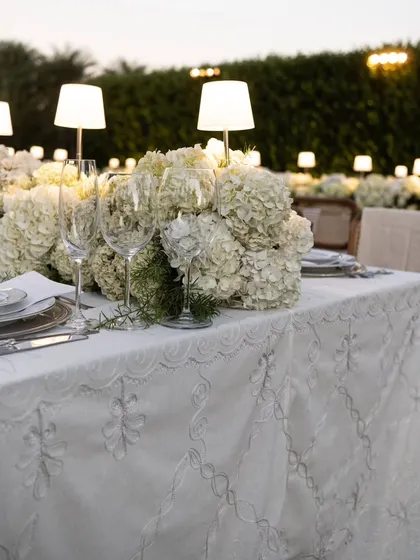 The edge of a dining table, showing the intricate embroidery on the white tablecloth and a lush centerpiece of white hydrangeas. Every detail is chosen to exude elegance.
