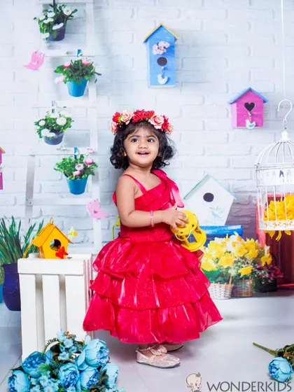 A toddler in a beautiful red dress, surrounded by birdhouses and flowers. The bright colors and her sweet smile make this a very cheerful and charming portrait.