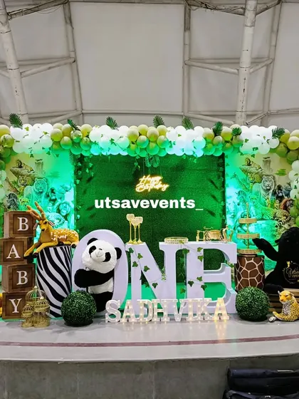 A fun jungle-themed first birthday with a green-lit backdrop. The setup includes large 'ONE' letters, various animal props, and a green and white balloon arch.