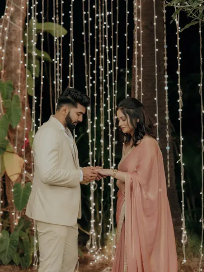 The ring exchange moment during an intimate engagement ceremony, set against a curtain of warm fairy lights.