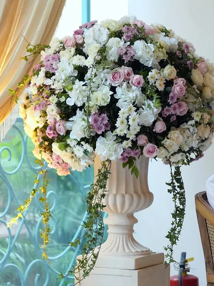 A large, classic urn filled with an overflowing arrangement of white, pink, and lilac flowers. This kind of statement piece is perfect for entrances or as a focal point in a large room.