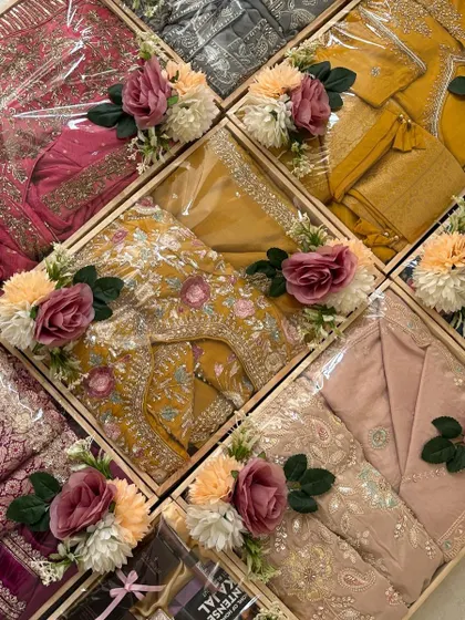 A beautiful array of wedding gifts packed in coordinated pinewood trays. Each gift, from sarees to suit sets, is wrapped to perfection and adorned with a matching floral arrangement of pink and peach roses.