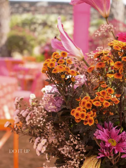 A close-up of a floral arrangement from a 'Rangilo Rajasthan' themed event. The mix of orange, pink, and purple flowers creates a beautiful, multi-tonal look.