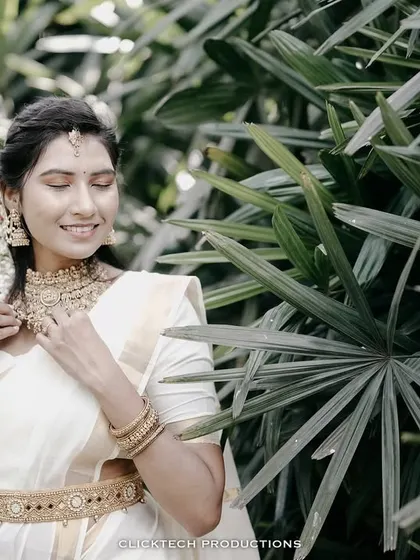 A beautiful solo portrait of the bride-to-be in a traditional white and gold saree, surrounded by lush green foliage.