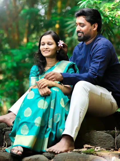 A sweet pre-wedding moment where the groom places a flower in the bride's hair as they sit together in a natural, green setting.
