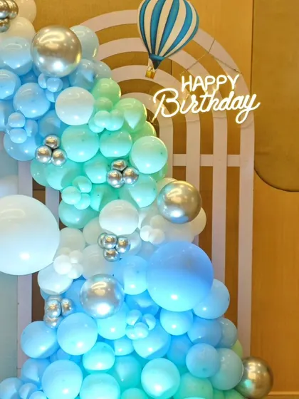 A hot air balloon-themed birthday decoration, featuring a beautiful balloon garland in shades of blue and white with silver accents and a 'Happy Birthday' neon sign.