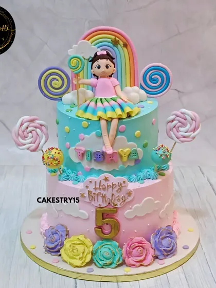 A magical two-tier Candyland rainbow cake for a fifth birthday. This black forest cake features a doll topper, colorful lollipops, and multi-colored roses against a pastel rainbow background.