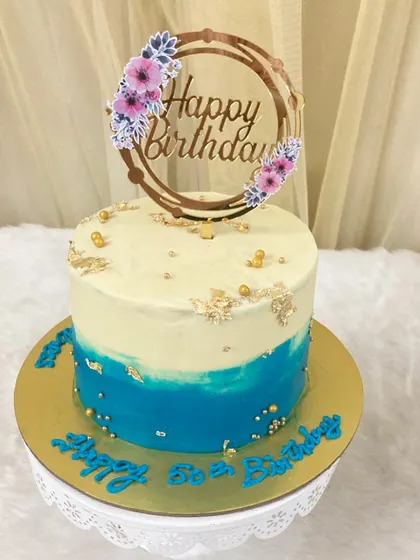 A simple but elegant buttercream cake in shades of white and blue, decorated with gold pearls and flakes for a 50th birthday.