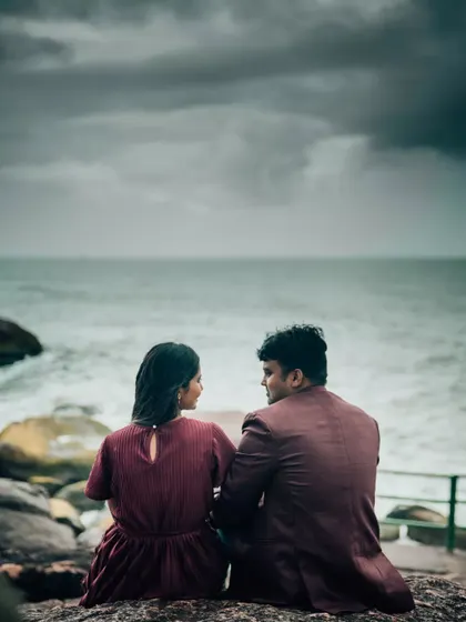 A couple sits on the rocks, looking out at the ocean on a dramatic, cloudy day. A beautiful, moody pre-wedding photograph.