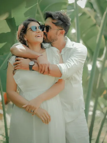 A fun and carefree moment captured against a backdrop of lush tropical leaves. The couple, dressed in matching white, shares a laugh, showcasing the relaxed and joyful side of their relationship.