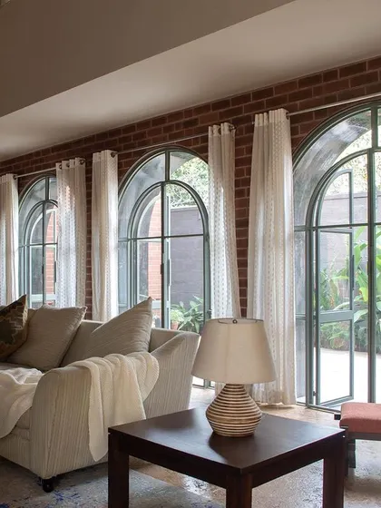 A series of arched French doors lines the living room, opening up to a lush green courtyard. Sheer white curtains soften the light and add a touch of elegance to the space.