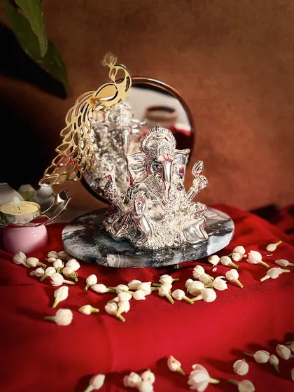 This stunning Ganesha idol combines divine artistry with reflective beauty. The silver-plated form is set against a mirror backdrop adorned with a golden peacock, all resting on a sophisticated grey marble base.