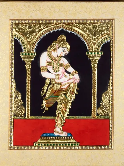 A framed Tanjore painting of a dancer, shot to highlight the intricate gold work and vibrant colors against the black background.