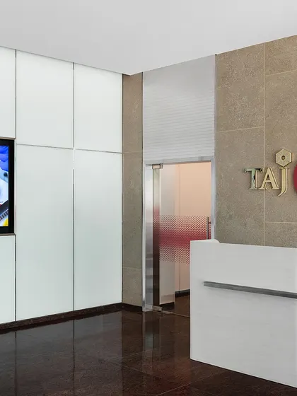 The informal reception area at the top of the stairs in 'The High-Flying Office'. A clean, modern reception desk is set against a stone wall with the TajSATS logo, creating a professional and welcoming first point of contact.