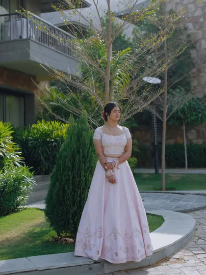 A serene portrait of the bride in a light pink lehenga, standing in a garden with her eyes closed, as if soaking in the moment.