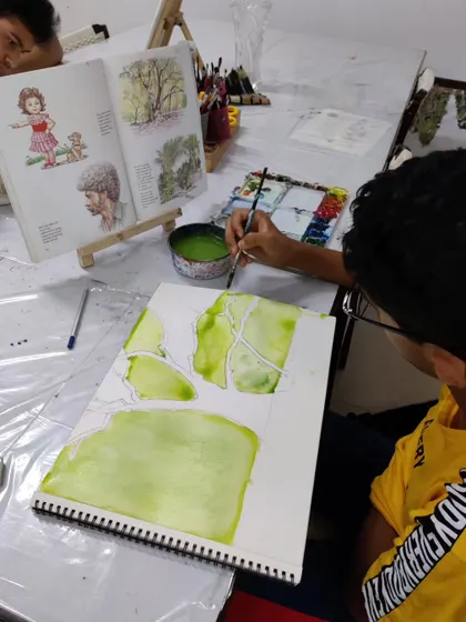 A student begins a tree study by applying a light green wash of watercolor, establishing the base layer for the foliage.