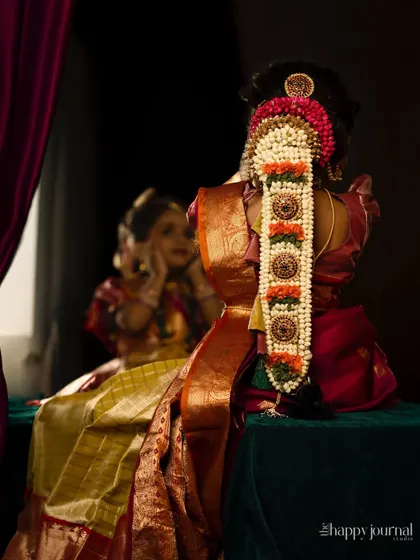 A girl in a traditional silk lehenga, her back to the camera, showing off the intricate 'moggina jade' floral hair arrangement.
