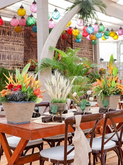 A dining table set for guests, featuring exotic floral centerpieces, woven placemats, and green glassware. The artificial palm tree adds to the immersive tropical atmosphere.