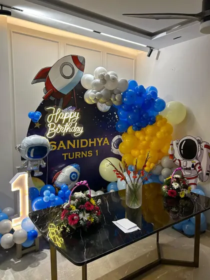 A compact space theme decoration for a first birthday, perfect for home setups. It includes a rocket and astronaut backdrop, a neon sign, and a light-up number.