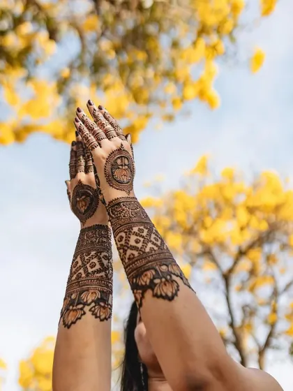 A beautiful shot of henna-adorned arms raised against a backdrop of yellow flowers. The design features lotus mandalas and geometric patterns.