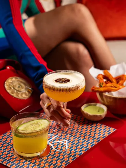 A vibrant scene at Lyla Bombay with two distinct cocktails and churros. The bright colors and playful composition create a fun and energetic vibe.