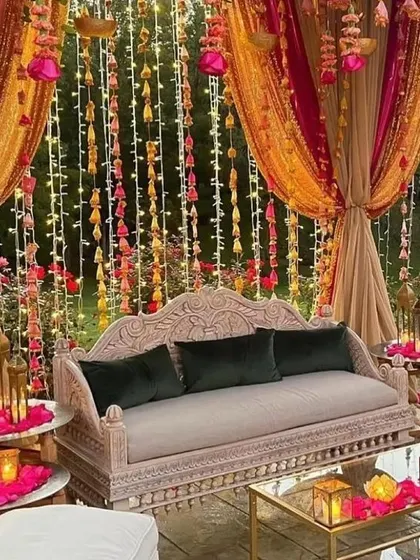 An intimate and ornate seating area perfect for a Mehendi. The carved sofa, combined with hanging floral strings and lanterns, creates a cozy and luxurious feel.