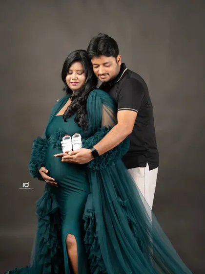 Holding tiny shoes, dreaming of tiny feet. This classic maternity pose with an elegant green gown against a dark, textured backdrop is both formal and deeply sentimental.
