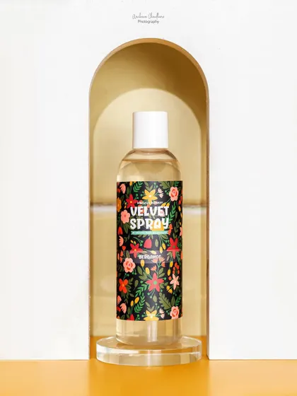 A clean, elegant shot of a body spray framed within a white arch prop. The simplicity of the composition puts all the focus on the product's vibrant label.