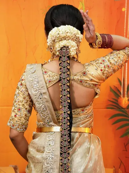 The back view of this bridal look highlights the stunning hairstyle and the detailed embroidery on the blouse. The cream and gold saree is complemented by a traditional floral braid.