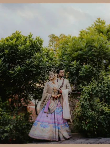 A regal portrait of a couple in exquisite designer outfits, standing amidst vibrant green trees. The bride's multi-colored lehenga by Tarun Tahiliani is a centerpiece of this elegant shot.