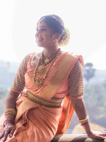 A serene bridal portrait. The bride, in her beautiful peach-colored saree, sits against a large window, bathed in soft, natural light.