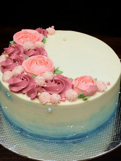 This milestone birthday cake features a blue watercolor effect on the bottom tier. The top is decorated with a crescent of hand-piped buttercream roses in shades of pink.