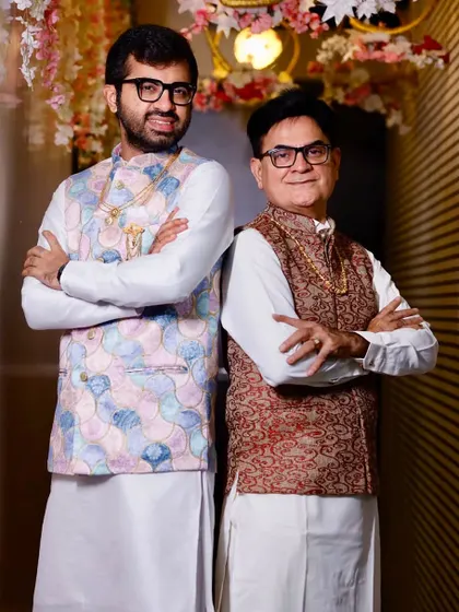 A father and son duo in custom outfits. The son wears a white kurta with a modern pastel-printed bandi jacket, while his father wears a classic red brocade jacket.