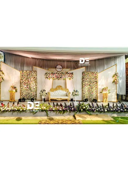 This wide shot showcases an outdoor wedding stage with elegant white and gold paneling and dense floral arrangements. The design creates a formal and sophisticated focal point in a natural setting.