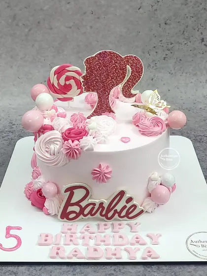 A pretty pink Barbie cake. The design includes a glittery Barbie silhouette, lollipops, and beautiful pink and white buttercream swirls.