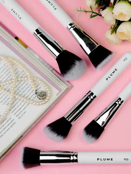 This flatlay of makeup brushes on a magazine, surrounded by pearls and flowers, creates a chic, editorial feel. The soft pink background enhances the feminine and sophisticated aesthetic of the products.