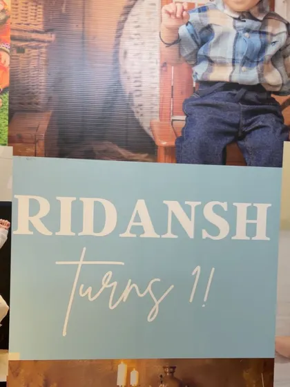 A close-up of the personalized "Ridansh Turns 1" sign, which serves as the centerpiece for the photo collage backdrop.